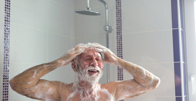 An older man smiles under a shower, covered in soap, washing with both hands. Light tiles and modern showerhead visible.