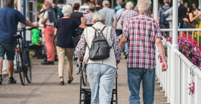 An elderly couple walks on a busy street; the woman uses a walker, the man wears a plaid shirt, and colorful flowers line their path.