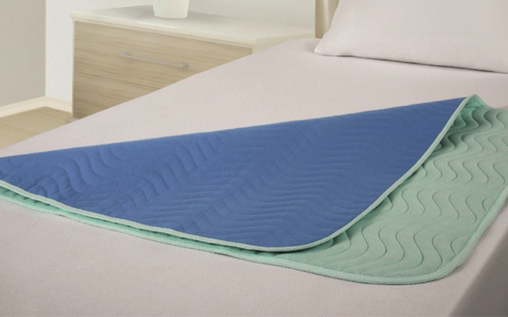 A bed has a blue and green mattress protector folded back, showing both sides. It has a wavy texture. A light nightstand is behind it.
