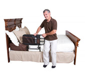 Man holding Stander Adjustable Bed Rail with support bar attached to side of wooden bed, in use, front view.