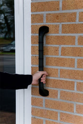 Hand holding a straight outdoor grab bar with anti-slip grip, fixed vertically to a brick wall near a glass door, in use.