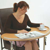 Cefndy Kidney Table with large adjustable surface, shown in use by woman sitting, holding a cup and reading a magazine, front view.