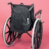Black wheelchair bag with side pocket holding a water bottle, attached to wheelchair, pink background, side view.