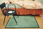 Dark grey waterproof mat under a commode chair next to a bed with floral bedspread and red skirt, in use view.
