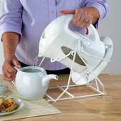 NRS Cordless Kettle Tipper with base clamps tilting a kettle to pour hot water into a teapot on a wooden table, front angle in use.