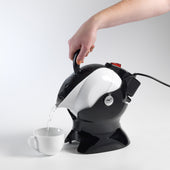 Hand pouring hot water from black and white Uccello Kettle into a white cup on a white surface, in use.