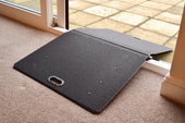 Portable black folding door threshold ramp, bridges carpet and patio tiles, shown unfolded, angled view.