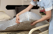 NRS waterproof fitted mattress cover being placed on hospital bed, chair and flowers in background, in use view.