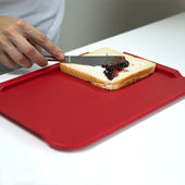 Hand spreading dark jam on bread with a knife, ORNAMIN large red non-slip cutting board in use, overhead view.