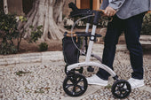 Person wearing jeans and white shoes using Saljol Allround Rollator outdoors on a stone path near a tree, in use view.