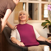 Older woman wearing a burgundy tabard style adult bib, orchid in background, cosy bright room, front view.