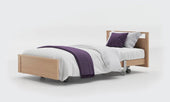 Oak low footboard profiling bed with wheels, white bedding, purple throw and matching pillow, side view.