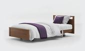 Oak profiling bed with low footboard, wheels, white bedding, and purple blanket and pillow, side view.