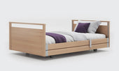 Oak bariatric profiling bed with wheels, white and purple bedding on a plain white background, front view.