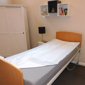 Single bed with satin SIBA sheets next to a white wardrobe, lamp, and shelves holding books and toys, side view.