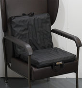 Black Harley 2-Way Sculptured Support Cushion with handles on a brown armchair, white wall background, front view.