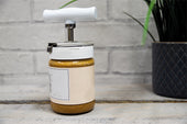 Aidapt jar opener with white handle on a table, plant blurred in background, front view.