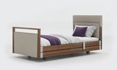 Beige upholstered profiling bed with wooden frame, white bedding and purple accents, front view.