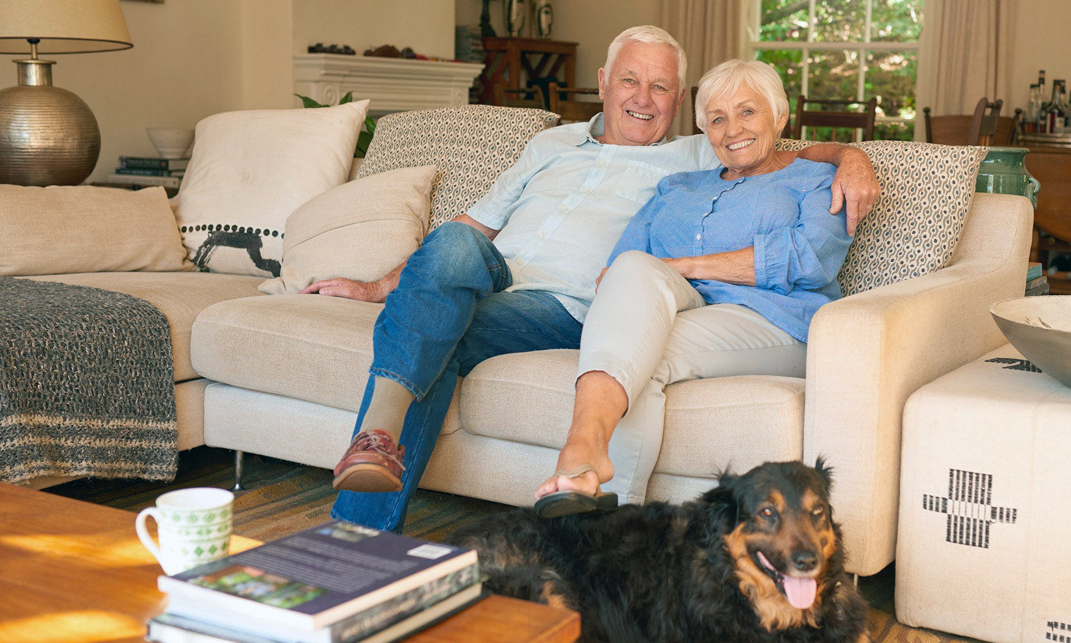 Older couple sitting together on a beige sofa, smiling and relaxed in a cosy living room. A black and brown dog lies contentedly on the floor in front of them.