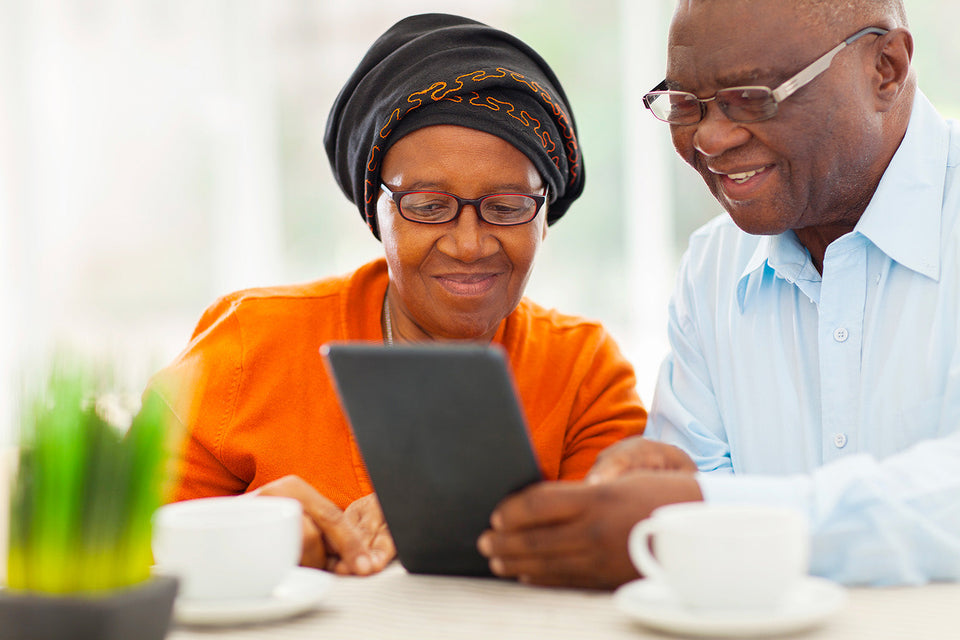 An elderly couple sits together at a table, smiling and looking at a tablet device. The woman is wearing an orange outfit with a black headscarf and glasses, while the man is in a light blue shirt and glasses. There are two white coffee cups and a small green plant on the table in front of them.