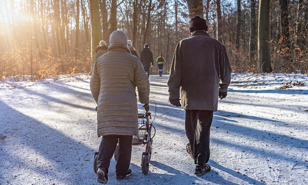 Elderly couple walking along a snowy woodland path in winter, with one person using a rollator walker as others walk ahead in the distance.