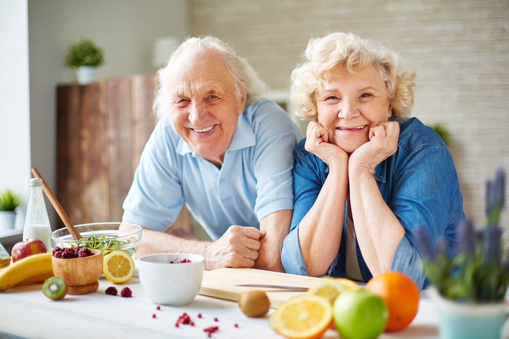 An elderly couple smiles, preparing food with fresh fruits like oranges and berries in a kitchen. Theyre wearing casual blue clothes.