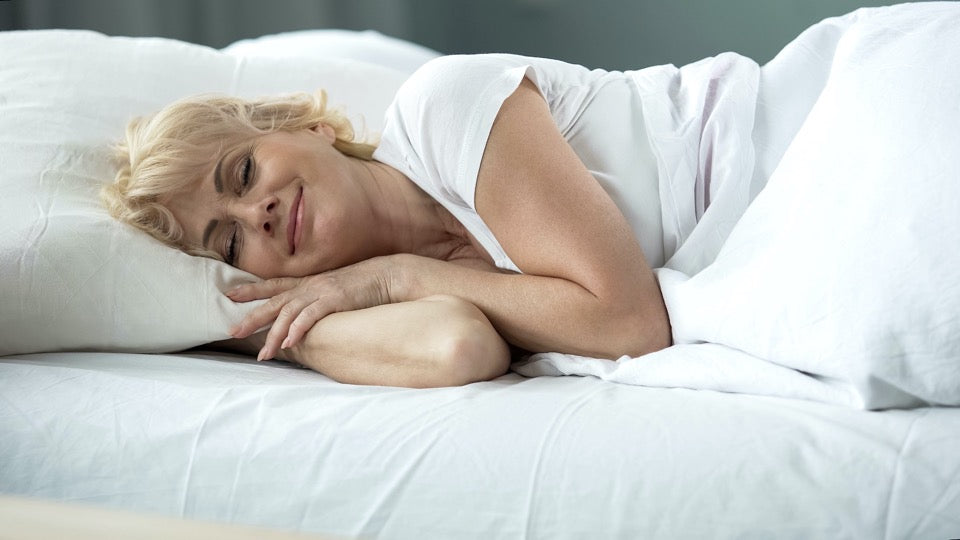 A woman sleeps peacefully on her side in a white bed, wearing a white shirt. The surroundings are all white, creating a calm scene.