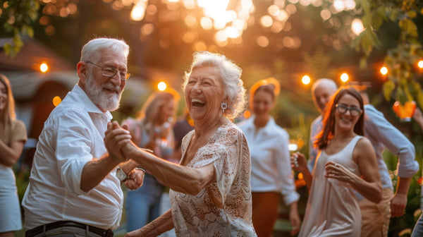 An elderly couple dances happily outdoors at sunset, surrounded by smiling people in a garden. Warm lights create a festive atmosphere.