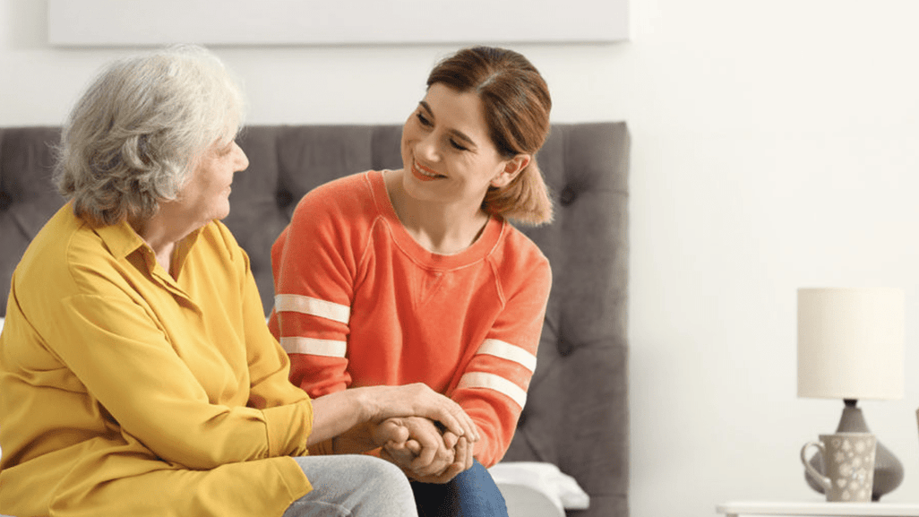 A young lady in an orange sweater and an older lady in a yellow blouse hold hands on a bed, smiling warmly at each other.