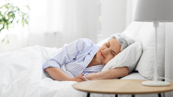 An older adult sleeps peacefully in bed, wearing light blue striped pajamas. A plant and wooden table are visible in the softly lit room. Image