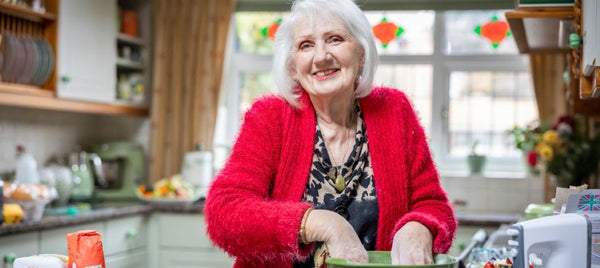 An elderly woman in a red cardigan smiles while baking, mixing ingredients in a green bowl in a sunlit kitchen. Image