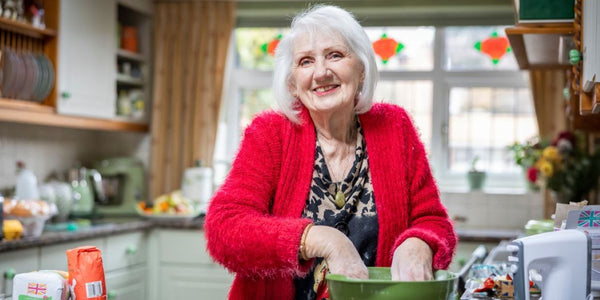 An elderly woman in a red cardigan smiles while baking, mixing ingredients in a green bowl in a sunlit kitchen.