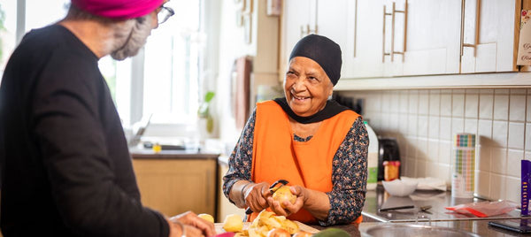 An elderly woman in an orange apron peels vegetables, smiling. A man in a pink turban slices food nearby amidst kitchen tools and ingredients. Image