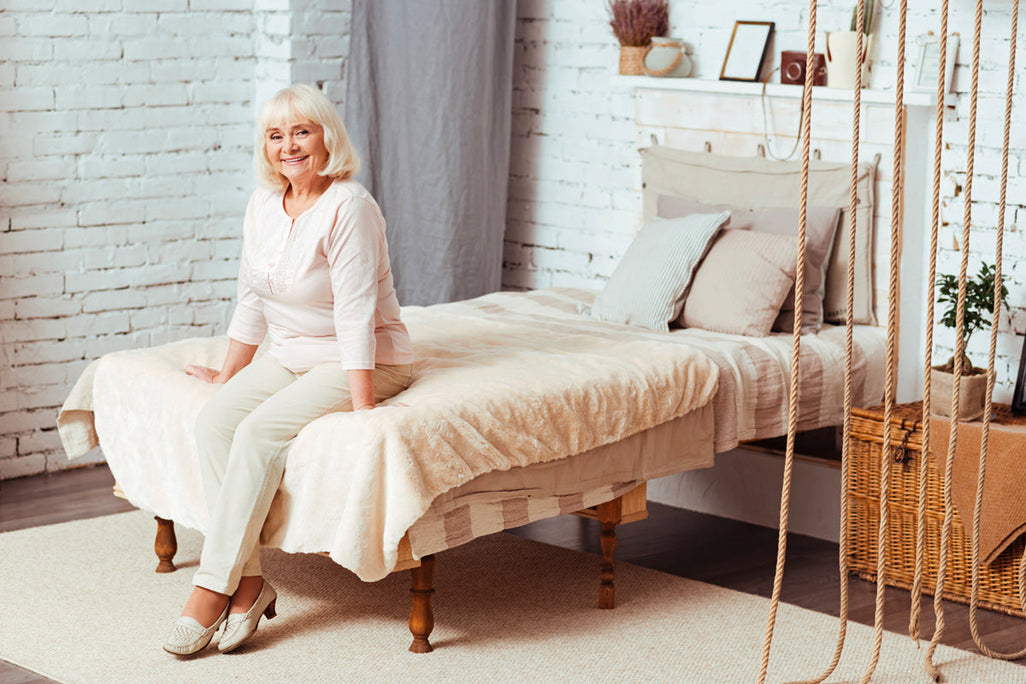 An elderly woman sits on a bed in a cozy room with white brick walls, neutral bedding, a woven basket, and shelf decorations.