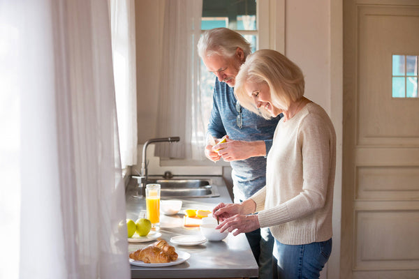 An older couple enjoys breakfast in a bright kitchen with fruit, croissants, and juice on the counter. White curtains frame the window.