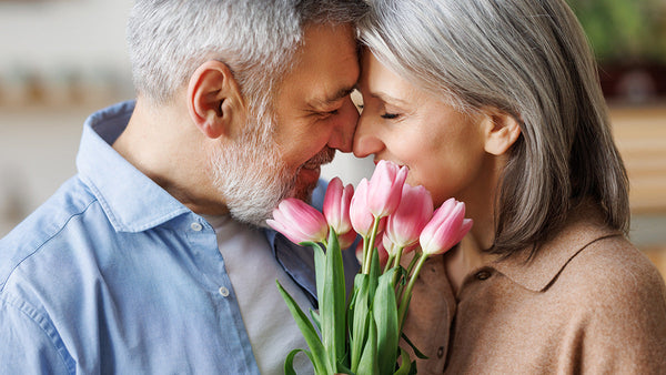 An elderly couple smiles with their foreheads touching, holding a bouquet of pink tulips, looking happy and content.