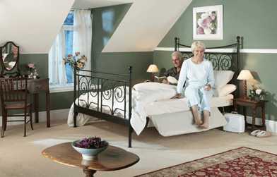 A cozy bedroom has green walls, a large bed with an elderly woman and a man sitting. A wooden table and lamps add warmth.
