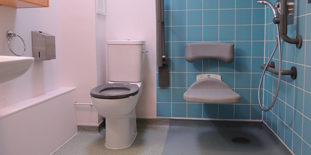 A bathroom with a white toilet, gray grab bars, a wall-mounted shower; light blue tiled shower area with a foldable gray seat.