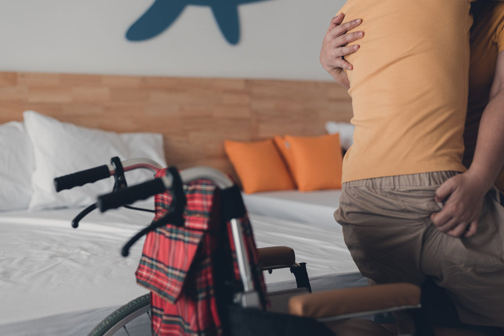 A person is being helped onto a bed with someone supporting them. A wheelchair and plaid blanket are nearby; orange pillows are on the bed.