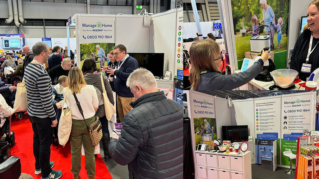 People stand at a booth labeled Manage At Home with posters, medical equipment, and lively conversations in a busy exhibit.