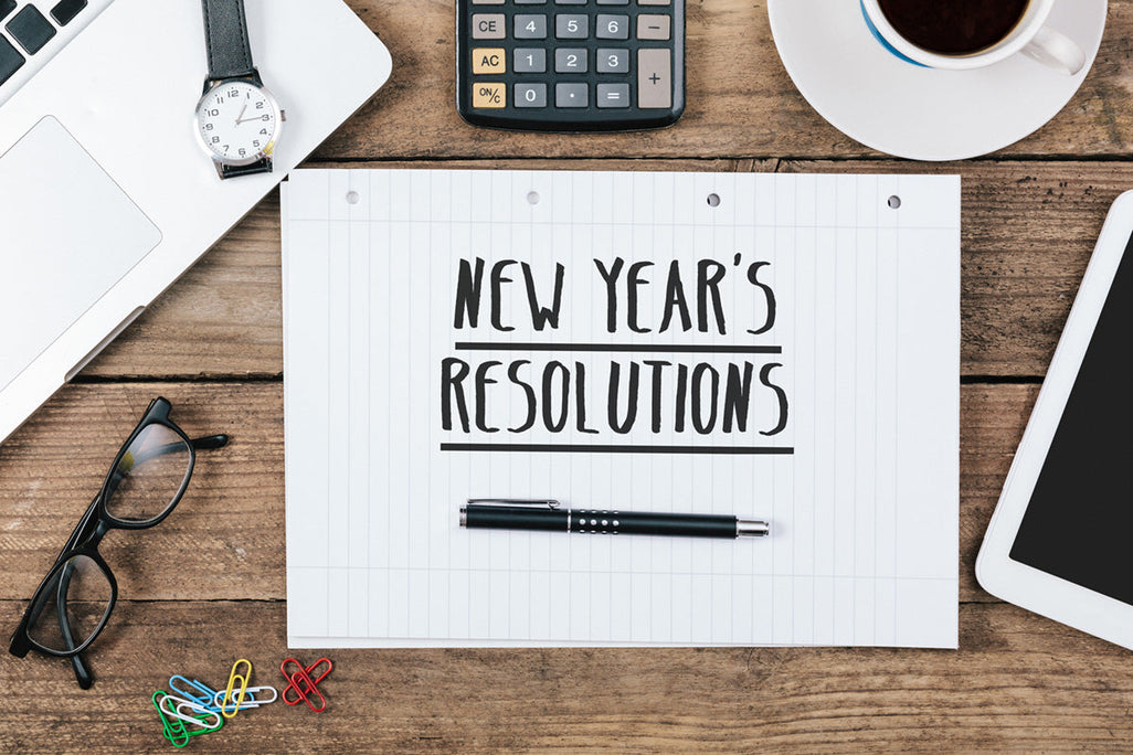 A wooden desk with a New Years Resolutions paper, pen, glasses, colorful paperclips, a laptop, calculator, tablet, and coffee cup.