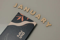 A 2025 calendar with a dark cover shows abstract orange and white leaves on a gray surface, with JANUARY in wooden letters above.