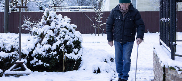 Older man walking carefully along a snowy garden path using a walking stick, with winter conditions around his home. Image