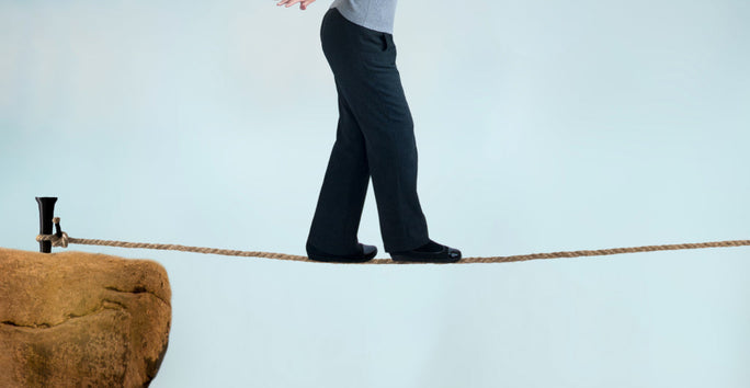 A person in black pants and shoes balances on a tightrope between a rock and an anchor, against a light blue sky.
