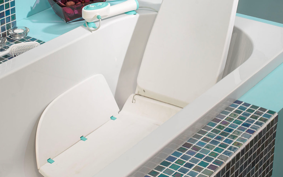 A white bathtub with a bath lift chair and blue-white tiles. A handheld shower head is next to a basket of various items.