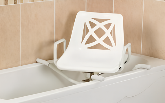A white bath chair with geometric cut-outs sits in a bathtub. It has armrests, set against beige tiled walls for easy tub access.