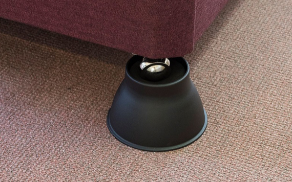 A black riser is placed under the leg of a red sofa on a carpeted floor, elevating the furniture for added height.