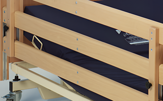 Close-up of a wooden hospital bed rail and blue mattress, with an ID tag visible. The bed is on wheels, and the rail is fastened with screws.