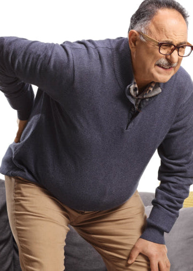 A man in glasses stands up from a couch, holding his lower back with a pained expression. The couch has yellow and gray cushions. Image