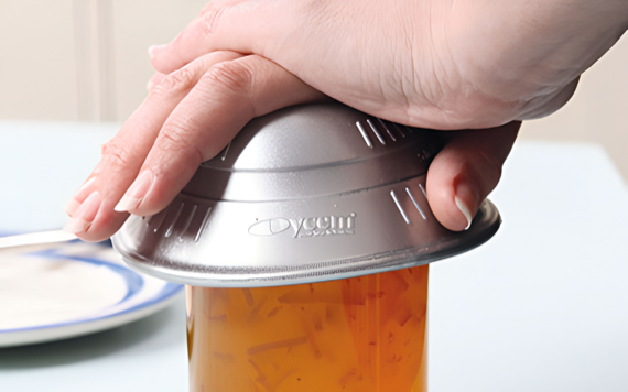 A hand securely holds a metal lid on a glass jar with orange liquid and chunks, on a light blue surface near a white plate with blue rim.